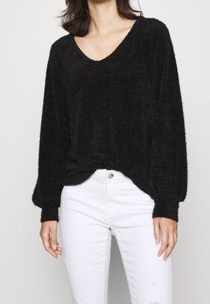 Femme portant un pull noir duveteux à col en V avec des manches bouffantes et un jean blanc taille haute sur un fond uni.