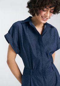 Granatowy kombinezon z krótkim rękawem, wykonany z lekkiego, teksturowanego materiału. Posiada kołnierzyk oraz dopasowaną talię z plisowanym detalem.