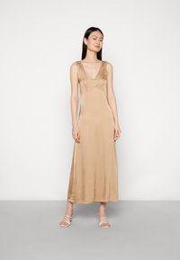 Vestido maxi em cetim de cor castanho claro com decote em V, corpete justo e saia fluida. A modelo usa sandálias brancas de tiras.