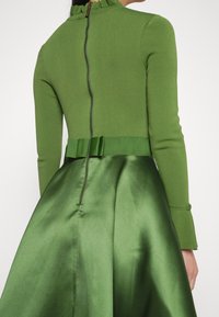 Vestido verde con un top ajustado de manga larga y cuello alto, con un borde arrugado, y una falda de satén con una silueta voluminosa y cremallera oculta.