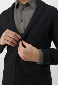 Schwarzer strukturierten Cardigan mit einem Schalkragen, vorne mit Knöpfen und zwei Taschen, getragen über einem grauen Hemd mit Knöpfen.