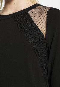 Musta paita, jossa on verkko- ja pitsikoristeet hartioilla, sileä kangas ja pyöreä kaula-aukko.