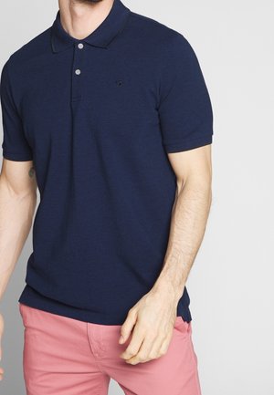 Man draagt een marineblauw poloshirt met korte mouwen en twee knopen, gecombineerd met een roze broek, tegen een effen lichte achtergrond.