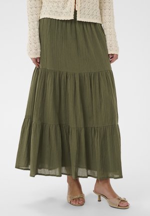 Žena v krémovom pletenom cardigane, olivovo zelenej viacvrstvovej maxi sukni a béžových otvorených topánkach na podpätku, stojaca na bielom pozadí.