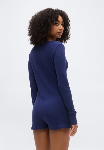 Γυναίκα με σγουρά μαλλιά, φορώντας μπλε σκούρο ολόσωμο φόρεμα με μακριά μανίκια, κοιτάζοντας πάνω από τον ώμο της σε απλό φωτεινό φόντο.