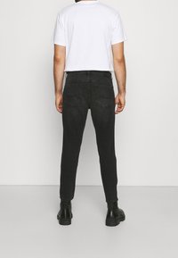 Muž v černých slim-fit džínech, bílém tričku s krátkým rukávem a černých botách, stojí čelem k jednoduché bílé zdi.