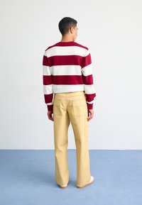 Suéter a rayas rojas y blancas de manga larga, combinado con pantalones amarillos con bolsillos traseros. La escena está ambientada sobre un suelo azul claro.
