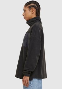 Pullover in pile nero con colletto alto, tasca frontale con zip in tessuto scuro, vestibilità comoda e polsini a costine. Indossato con jeans azzurri chiari.