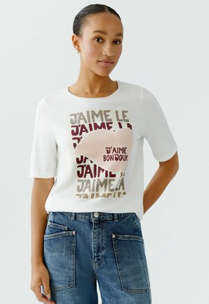 T-shirt blanc à manches courtes avec un motif de lèvres roses imprimé et du texte dans différentes polices et couleurs. Porté avec un pantalon en denim bleu foncé.
