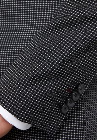 Rękaw czarno-białego żakietu w cekiny z teksturowanym materiałem, klapą typu notch oraz czterema czarnymi guzikami na mankiecie.