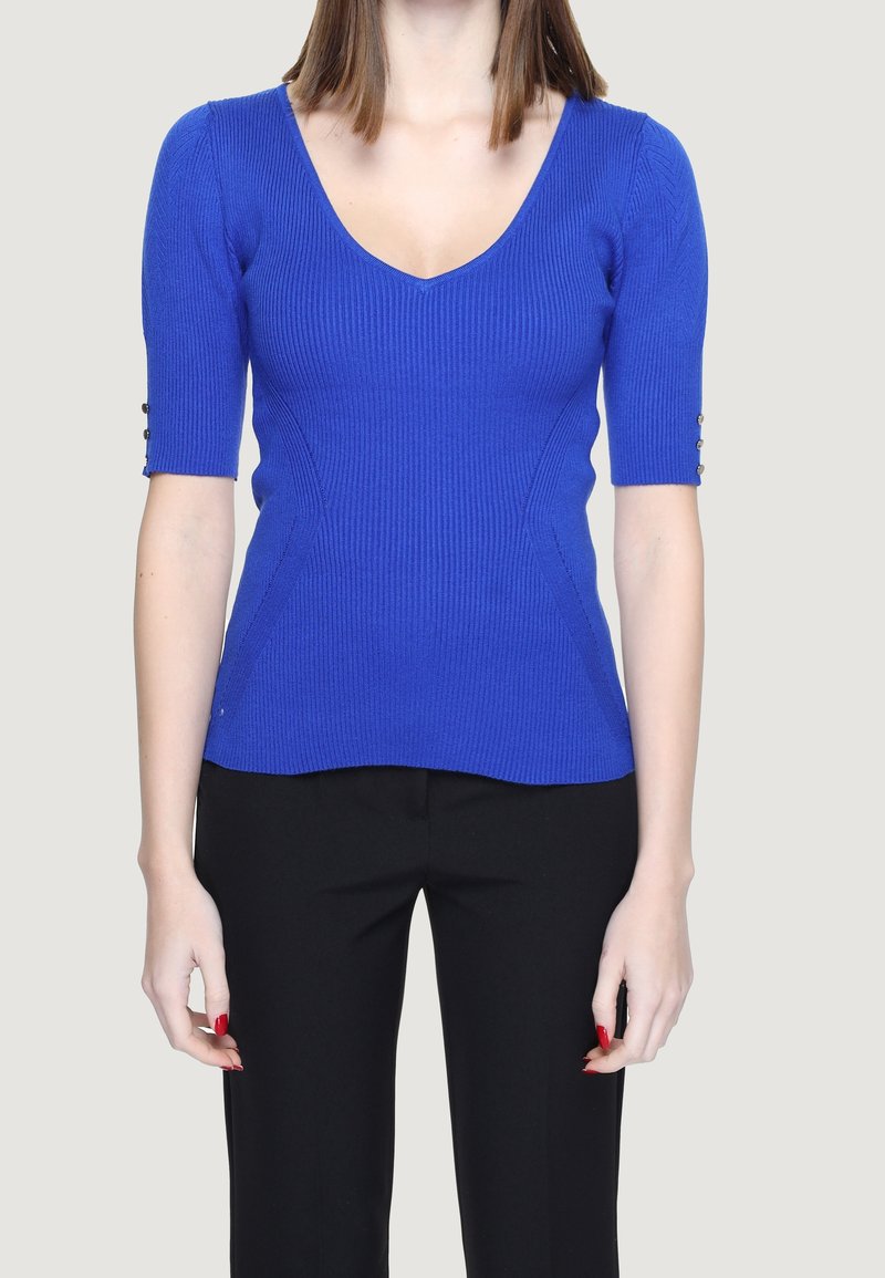 Femme portant un haut bleu ajusté côtelé à col en V avec des manches longueur coude et un pantalon noir, sur un fond clair uni.