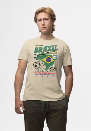 Jonge man draagt een beige T-shirt met Brazilië 1994 Wereldkampioenen voetbalontwerp, groene short en een neutrale gezichtsuitdrukking.