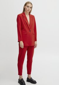 Blazer rosso e pantaloni tapered abbinati realizzati in tessuto liscio. Il blazer presenta un profondo scollo a V, chiusura con un bottone singolo e tasche laterali.