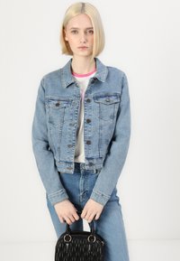 Chaqueta de mezclilla azul clara con un corte cropped, dos bolsillos en el pecho y cierre de botones. Combinada con una camiseta blanca y jeans azules, sosteniendo un bolso negro.