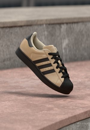 Beige suede sneaker with black rubber shell toe, three black stripes, and black laces. Flat sole, rounded shape, classic design.