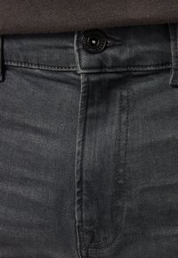 Gros plan de la ceinture d'un jean en denim noir avec fermeture par bouton et coutures visibles sous un haut gris foncé.