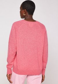 Una donna dai capelli corti indossa orecchini a cerchio dorati, un maglione rosa con texture e pantaloni a strisce rosa e bianchi, mostrata di spalle.