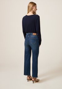 Mujer de pie de espaldas, vistiendo una blusa de manga larga azul oscuro, jeans de tiro alto azul y sandalias de tacón con estampado de serpiente sobre un fondo beige.