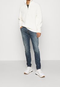 Weißer, strukturierter Strickpullover mit einem halben Reißverschluss, kombiniert mit dunkelblauen Jeans und weißen Sportschuhen mit hellbraunen Sohlen.