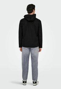 Giacca nera con cappuccio dalla texture liscia, vestibilità standard e taglio dritto. Abbinata a jeans grigio chiaro e sneakers nere.