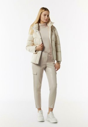 Helle Daunenjacke mit Reißverschluss, beigefarbenem Rollkragenpullover, eng anliegenden Kunstlederhosen und weißen Sneakern; glatte Texturen, lässige Passform.