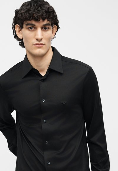Νέος άνδρας με σγουρά σκοτεινά μαλλιά, φορώντας μαύρο πουκάμισο με κουμπιά και μικρό κεντημένο λογότυπο στο στήθος, πάνω σε απλό φόντο.