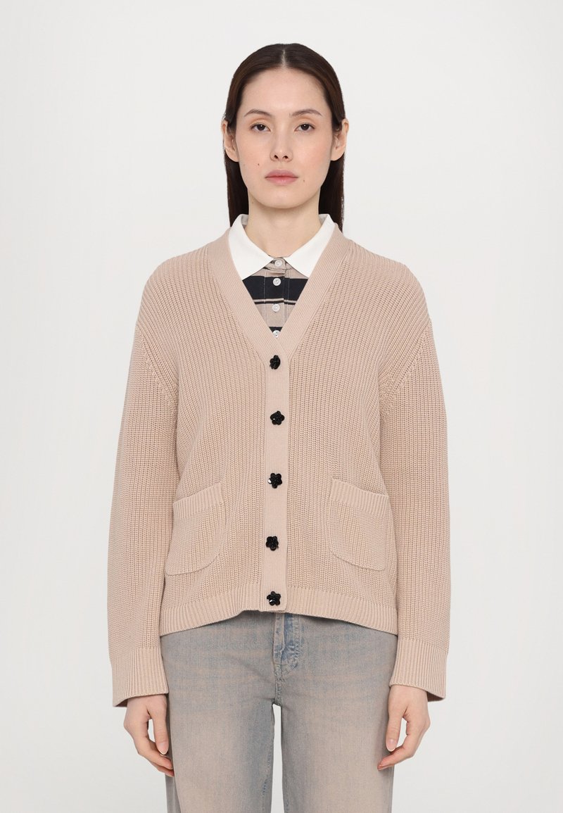 Μπεζ πλεκτό πουλόβερ με V-λαιμόκοψη, μαύρα κουμπιά και δύ�ο μπροστινές τσέπες. Φορεμένο πάνω από ένα καρό πουκάμισο με γιακά. Απαλή υφή.