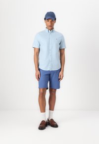 Chemise à manches courtes bleu clair avec des boutons, shorts bleus, mocassins marron et casquette de baseball bleue. Tissu lisse, coupe décontractée, design simple.