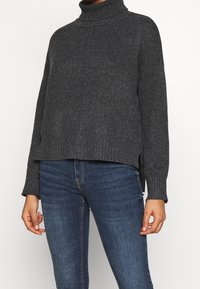 Suéter de cuello alto gris con un corte holgado, puños y dobladillo acanalados, aberturas laterales y una textura suave. Combinado con jeans azul oscuro.