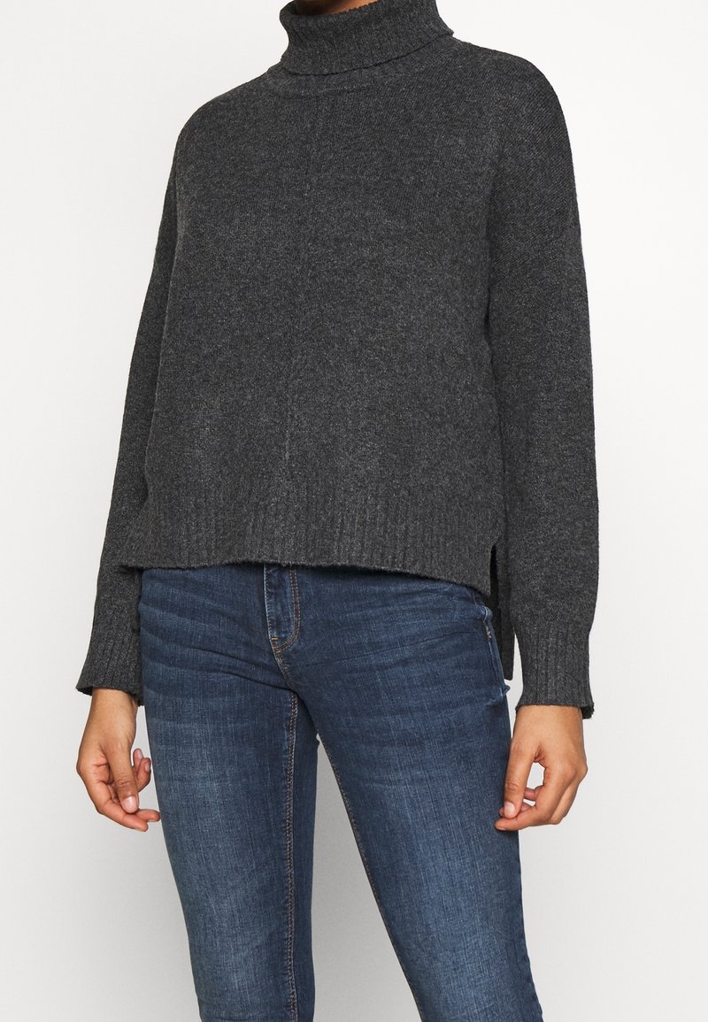 Suéter de cuello alto gris con un corte holgado, puños y dobladillo acanalados, aberturas laterales y una textura suave. Combinado con jeans azul oscuro.