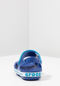 Sandalia de goma azul con correa trasera, que presenta perforaciones para ventilación y una banda blanca con la marca "crocs" en texto azul.