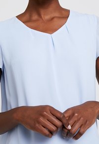 Light blue short-sleeve top with a V-neckline, featuring pleat detailing and smooth fabric. Hands are positioned at the waist.