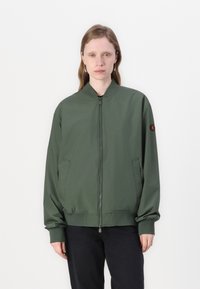 Groene bomberjack met een rits aan de voorkant, ribgebreide manchetten en twee zijzakken. Heeft een minimalistisch ontwerp en een ronde logo patch op de mouw.