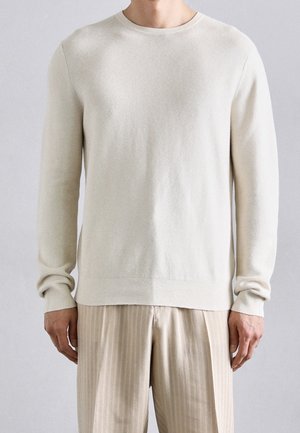 Homme portant un pull crème à manches longues et col rond, et un pantalon beige rayé, debout devant un fond clair uni.