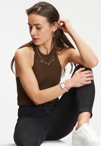 Braun gerippter Halsabschneider, geschichtete Halsketten mit rosa Akzenten, silbernes Armband, schwarze Jeans und weiße Sneakers. Glatte Texturen insgesamt.