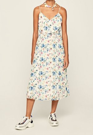 Femme portant une robe midi fleurie sans manches ceinturée, associée à des baskets blanches épaisses et un collier en coquillage.