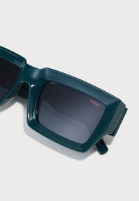 Lielas teal krāsas saulesbrilles ar taisnstūrveida objektīviem, ar gludu apdari un diskrētu "HUGO" logo sarkanā krāsā uz viena objektīva.