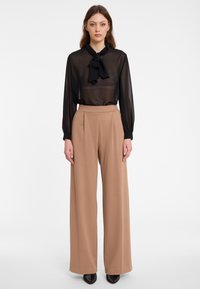 Zwarte doorschijnende blouse met een gestrikte strikdetail om de nek, gecombineerd met hoog-getailleerde, wijde, beige broeken. Soepele, lichte stof.