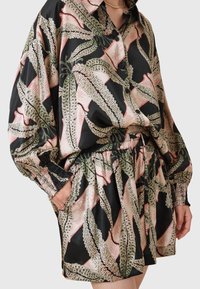 Femme portant une robe noire boutonnée avec des motifs de feuilles vertes et beiges, avec de longues manches bouffantes et une taille élastique avec cordon.