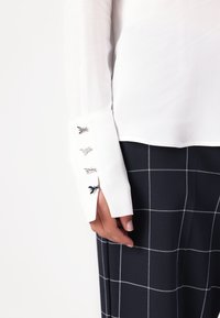 Camisa blanca de manga larga con puños con abertura y detalles decorativos en plateado, combinada con una falda de patrón a cuadros en azul marino. Textura de tela suave.