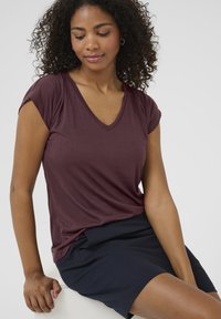 T-shirt bordeaux à col en V avec des manches courtes roulées, fabriqué à partir d'un tissu doux. Porté avec une jupe sombre, présenté sur un fond clair.