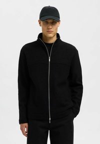 Svart zip-up jacka i texturerat tyg, med hög krage, en slät dragkedja och en avslappnad passform. Bärs tillsammans med en matchande svart keps.