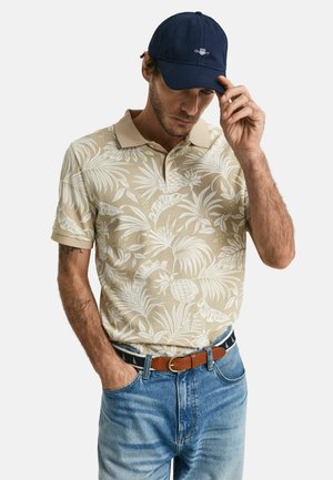 Beige polo shirt med vitt tropiskt blad- och ananasmönster, beige krage; mörkblå keps; blå jeans med brunt bälte, som visar ett avslappnat utseende.