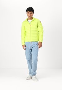 Bright yellow padded jacket with a zip front, high collar, and elastic cuffs, paired with light blue jeans and white sneakers.