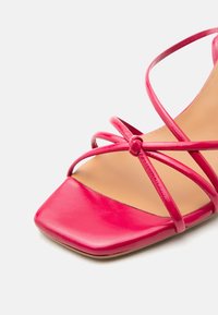 Κόκκινο σανδάλι ανοιχτού τύπου με τετράγωνη μπροστινή πλευρά και λεπτά διασταυρούμενα λουράκια πάνω από το πόδι μπροστά σε απλό λευκό φόντο.