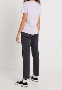 Camiseta de manga corta lila, vaqueros de tiro alto negros y zapatillas negras con suelas blancas. Cabello en ondas sueltas, visto desde atrás.