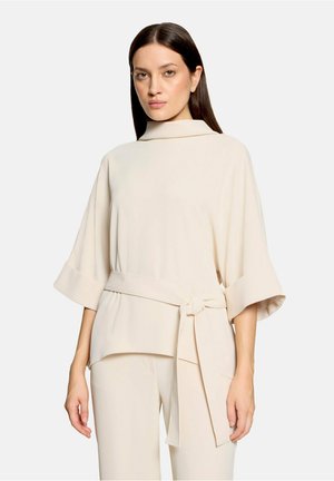 Femme aux cheveux raides et foncés portant un haut ample crème à manches larges et une ceinture en tissu nouée, assortie à un pantalon crème.
