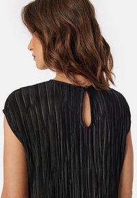 Czarny top z plisami i krótkimi rękawami, z zapięciem na plecach w kształcie otworu. Materiał ma teksturowany, zwiewny design.