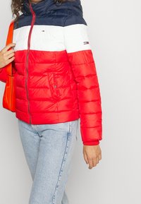 Kvinna i en röd, vit och marinblå puffarjacka med en röd dragkedja och ljusblå jeans, bärande en lysande orange handväska över axeln.