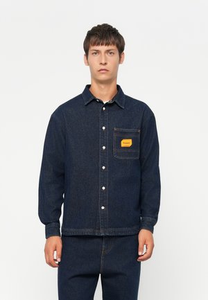Mørk denimskjorte med lange ermer, knaplukning og en brystlomme med en gul label. Glat tekstur med klassisk kravedesign.
